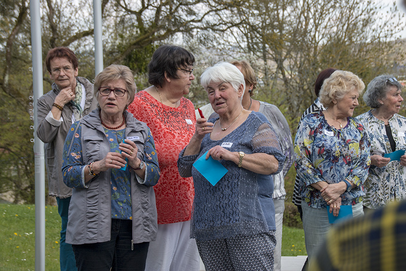 Eine Gruppe älterer Frauen steht auf einer Wiese in der Nähe der Hochschule Coburg und unterhält sich. Sie halten blaue Papiere in den Händen und tragen Namensschilder. Im Hintergrund sind Bäume zu sehen.