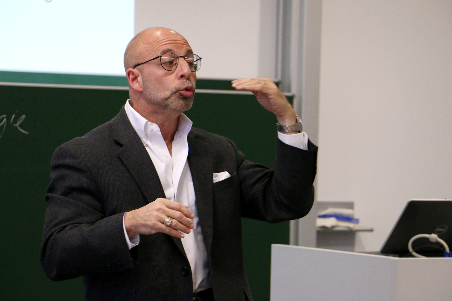 Ein kahlköpfiger Mann im Anzug gestikuliert, während er leidenschaftlich vor einem Hörsaal der Hochschule Coburg spricht. Auf dem Rednerpult in der Nähe steht ein Laptop, und im Hintergrund ist eine Tafel zu sehen, die der spannenden Vorlesung eine akademische Note verleiht.