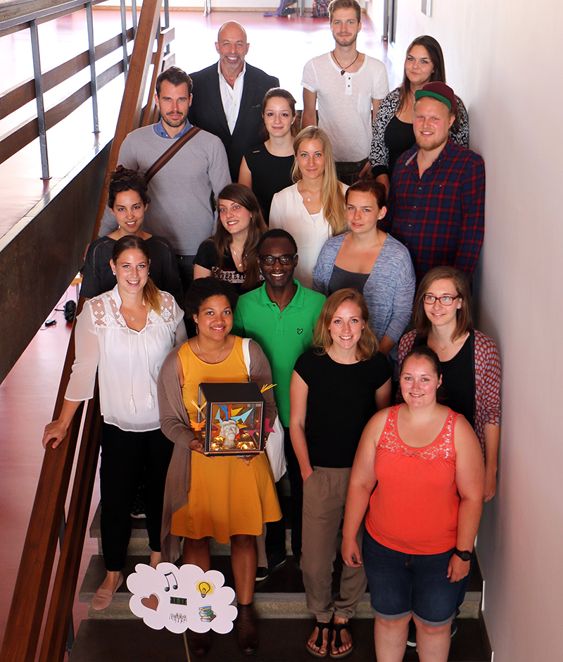 Eine bunt gemischte Gruppe von 16 Personen steht auf einer Treppe der Hochschule Coburg und posiert für ein Foto. Eine Person in der ersten Reihe hält ein gerahmtes Kunstwerk, während eine andere ein Schild mit Zeichnungen einer Wolke, einer Glühbirne und Musiknoten hält. Die Kulisse ist ein Flur mit Metallgeländern.