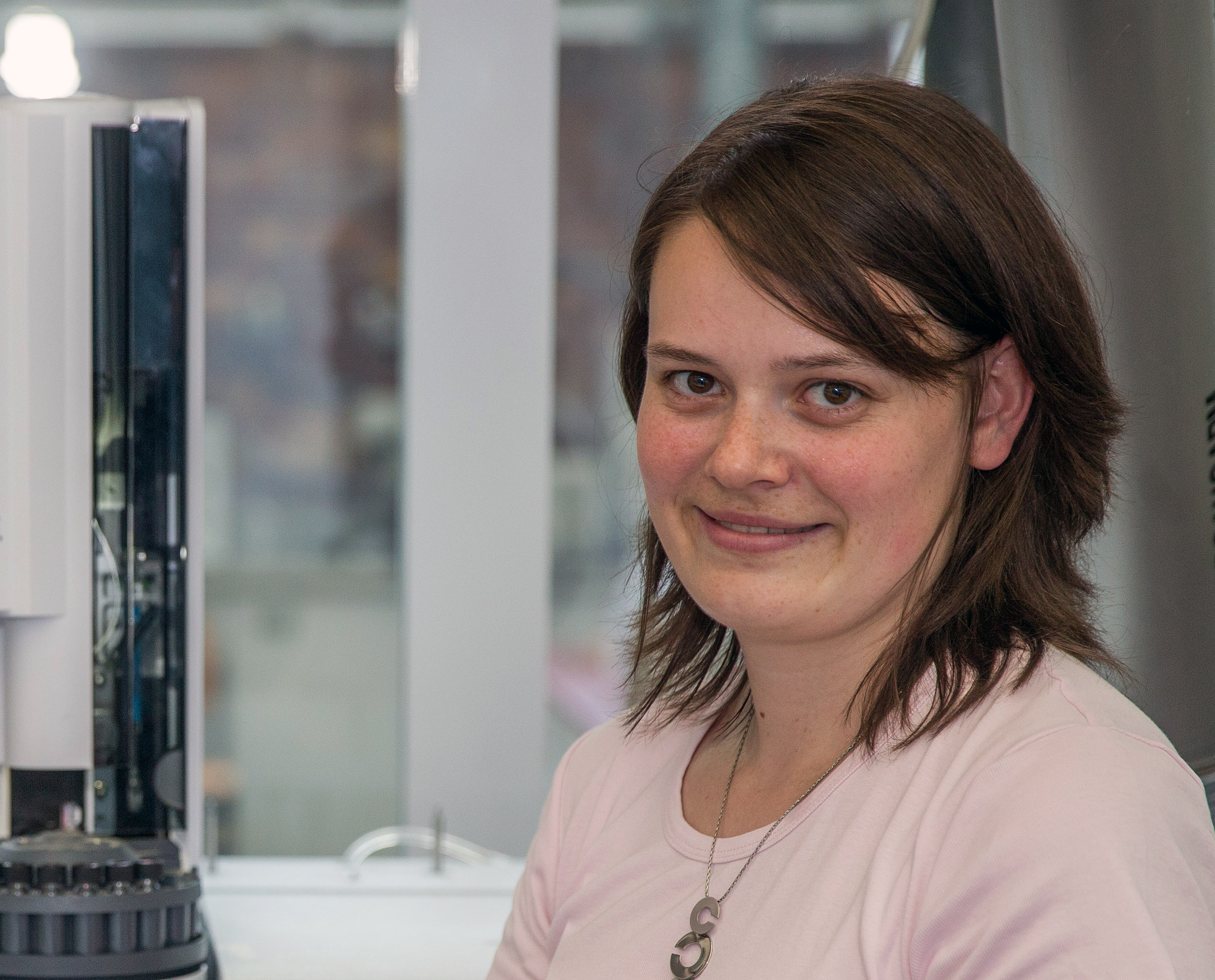 Eine Person mit schulterlangem braunem Haar, einem rosa Hemd und einer Halskette mit einem „S“-Anhänger lächelt in die Kamera. Sie steht in einem Labor der Hochschule Coburg neben einigen Maschinen.