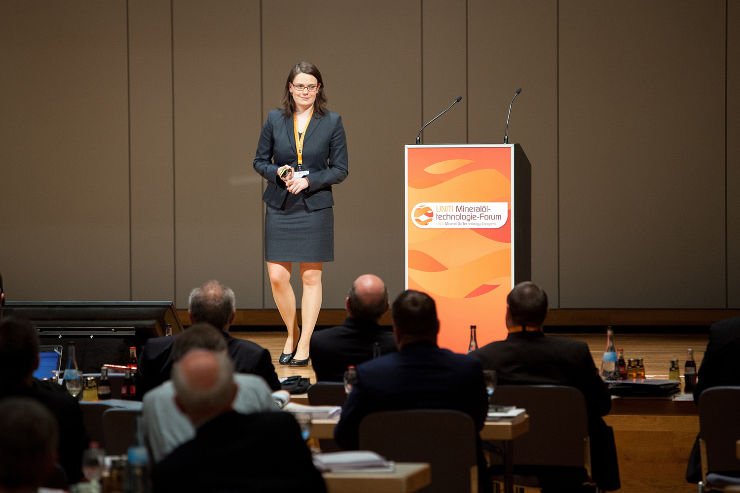 Eine Frau in Businesskleidung steht auf einer Bühne in der Nähe eines Podiums und hält einen Präsentationsklicker in der Hand. Ein Publikum aus sitzenden Personen beobachtet die Präsentation. Auf dem Podium hängt ein Schild mit der Aufschrift „UMT Mineralogische Technologien-Forum“, Mitveranstalter ist die Hochschule Coburg.