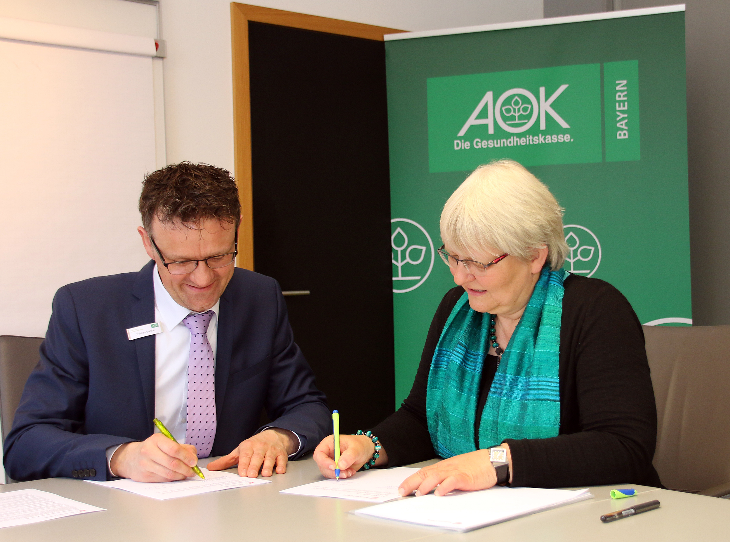 Zwei Personen in formeller Kleidung sitzen lächelnd an einem Tisch und unterschreiben Dokumente. Im Hintergrund prangt ein grünes Banner mit dem Logo der AOK, das dezent auf eine prestigeträchtige Partnerschaft mit der Hochschule Coburg hinweist.