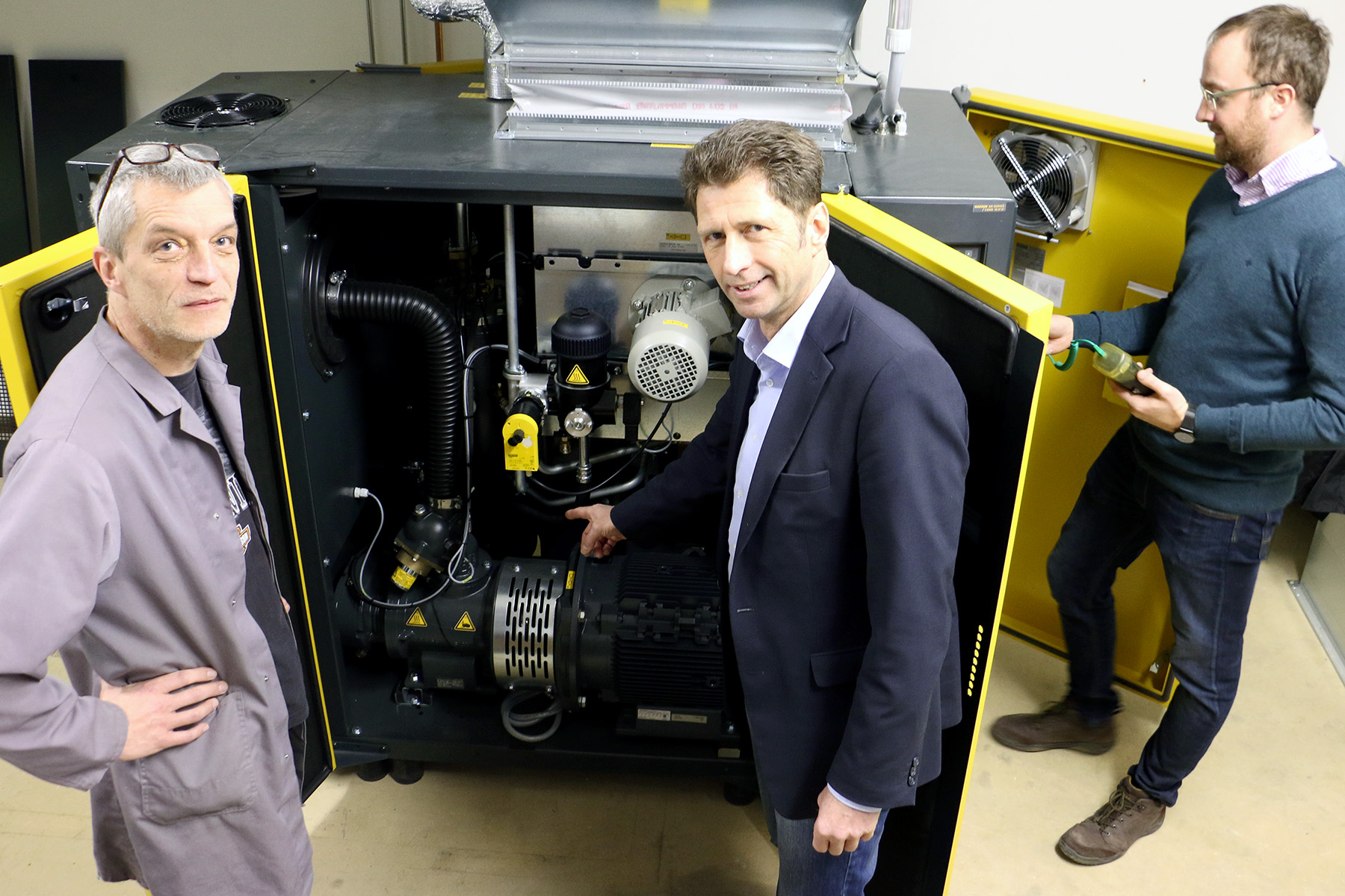 Drei Männer der Hochschule Coburg prüfen Maschinen in einem offenen Schrank. Ein Mann zeigt auf ein Bauteil, ein anderer hält ein Werkzeug und der dritte steht daneben. Der Schrank enthält verschiedene Rohre und mechanische Teile, was auf eine laufende Ingenieurszusammenarbeit schließen lässt.