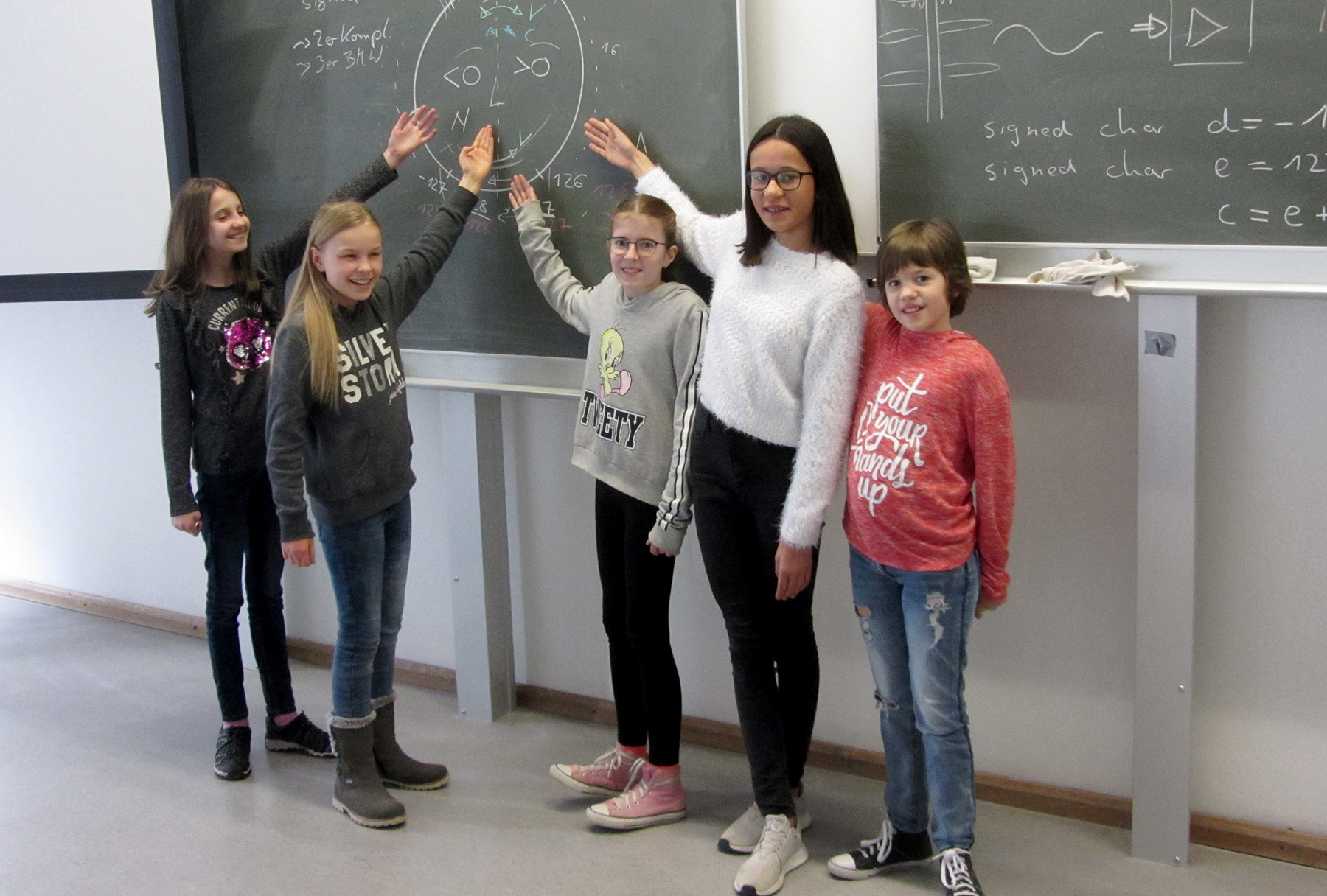 Fünf Studierende stehen an der Hochschule Coburg vor einer Tafel, lächeln und zeigen auf gezeichnete Diagramme und Gleichungen. Auf der Tafel sind Schleifen und Kodierungsbegriffe eingezeichnet, was auf eine Unterrichtsstunde zum Thema Programmieren hindeutet. Sie wirken in der Unterrichtssituation engagiert und kooperativ.