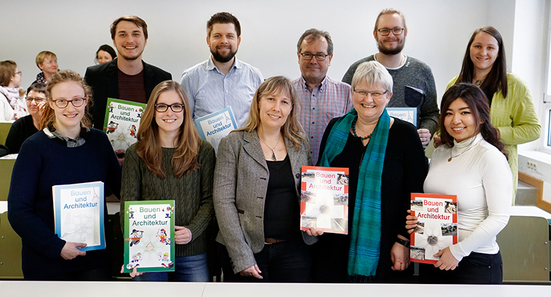 Eine Gruppe von zehn Personen steht in einem Hörsaal der Hochschule Coburg und lächelt in die Kamera. Sie halten Bücher mit dem Titel „Bauen und Architektur“ in den Händen. Im Hintergrund sitzen ein paar weitere Personen.