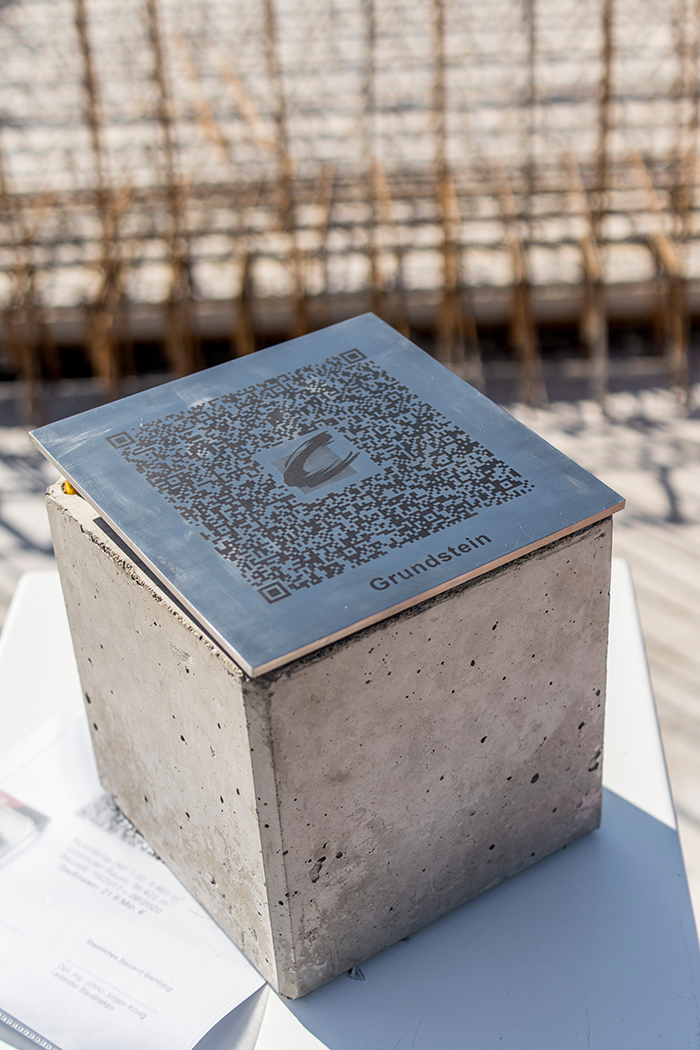 Ein Betonblock mit Metalldeckel zeigt einen auffälligen QR-Code und die Aufschrift „Grundstein“, was auf seine Bedeutung hinweist. Er steht auf einer hellen Oberfläche und daneben ist teilweise Papier zu sehen. Er gehört zur Vision der Hochschule Coburg. Der Hintergrund ist durch Baumaterialien verschwommen.