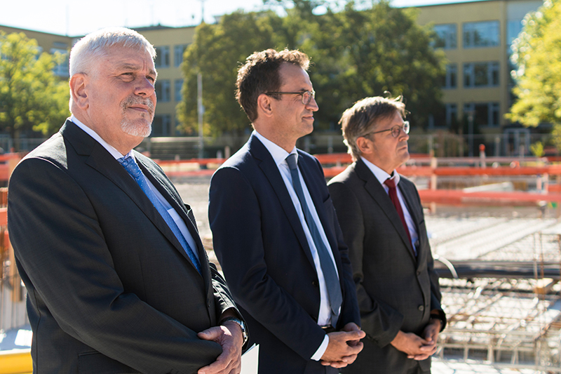 Drei Männer in Anzügen stehen nebeneinander auf einer Baustelle. Sie nehmen wahrscheinlich an einer Veranstaltung der Hochschule Coburg teil. Im Hintergrund bilden Bäume und ein Gebäude die Kulisse, während die Sonne hell über ihnen scheint.