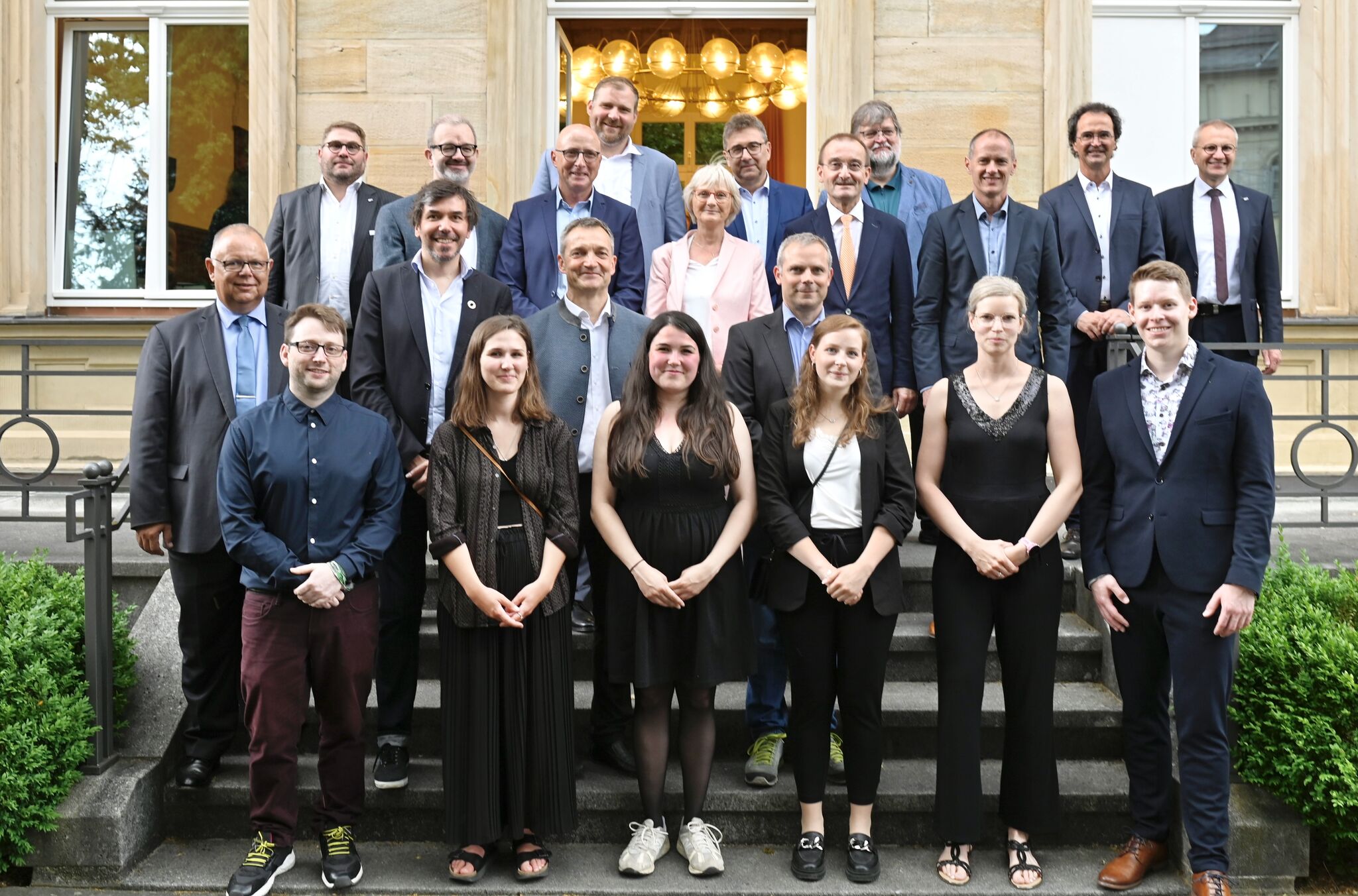 Eine Gruppe von Menschen, Männer und Frauen, steht auf der Treppe vor einem Gebäude der Hochschule Coburg. Einige sind formell gekleidet und tragen Anzüge, andere sind eher leger gekleidet. Sie stehen in mehreren Reihen, lächeln und posieren für das Foto.