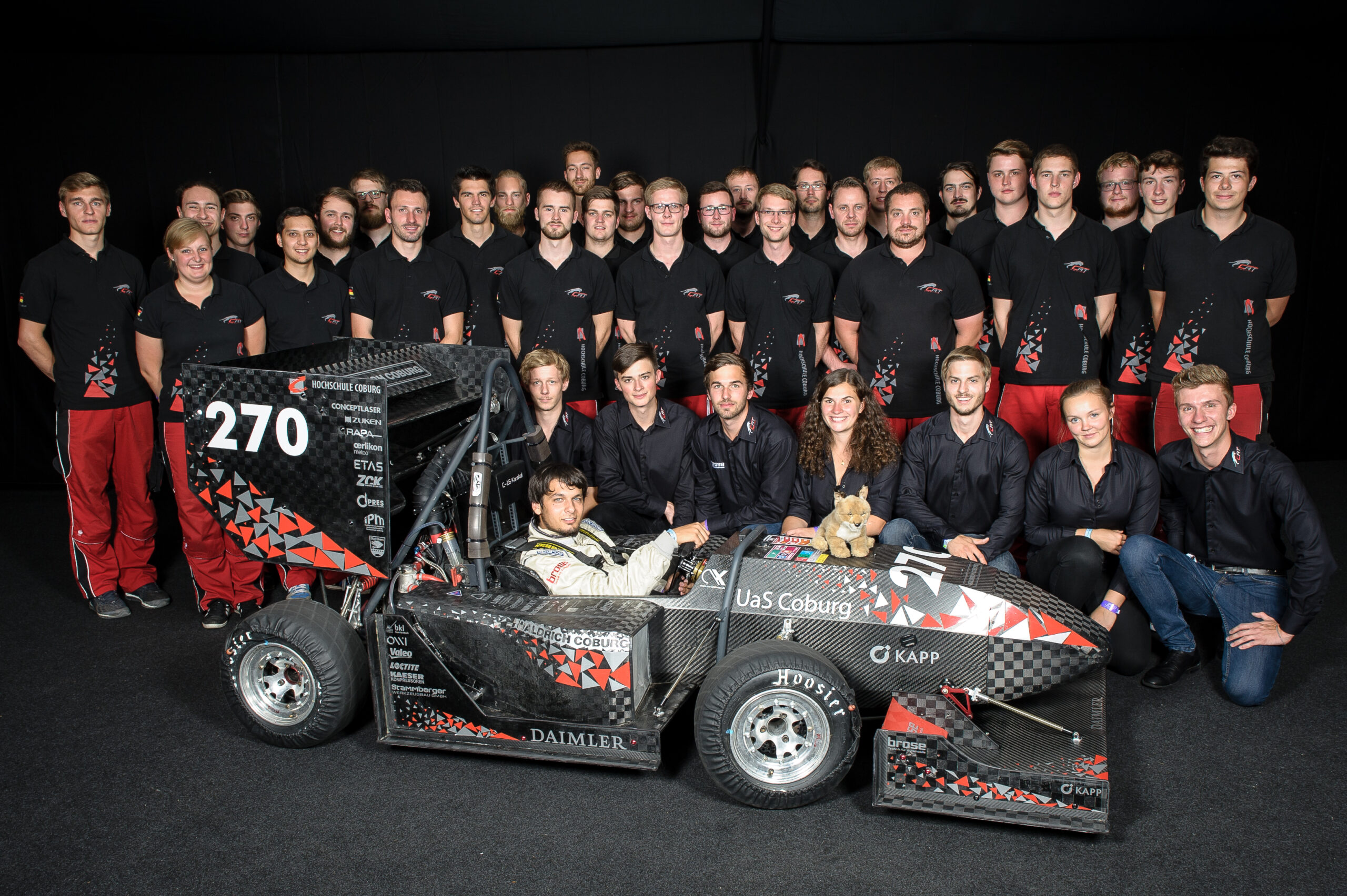 Eine Gruppe von 34 Personen in passenden Teamshirts der Hochschule Coburg posiert um einen Formel-Rennwagen. Das Auto ist schwarz mit geometrischem Muster und der Aufschrift "270". Im Auto sitzt ein Fahrer im Rennanzug, auf dem Gefährt sitzt ein Teddybär.