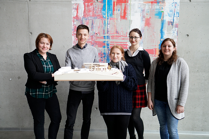 Fünf Menschen stehen lächelnd vor einem großen abstrakten Gemälde, leger gekleidet und mit einem Projekt in den Händen. Sie stehen vor einer Betonwand und verkörpern den kreativen Geist, der an der Hochschule Coburg entwickelt wurde.