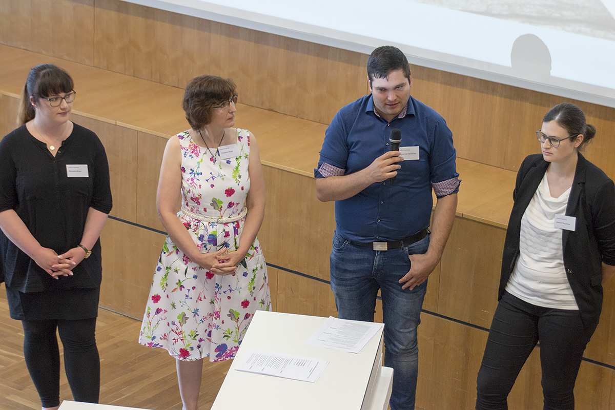 Vier Personen stehen in einem Hörsaal der Hochschule Coburg, zwei halten Mikrofone in der Hand. Drei sind dunkel gekleidet, eine sorgt mit einem geblümten Kleid für einen Farbtupfer. Sie scheinen in eine Diskussion oder einen Vortrag vertieft zu sein. Im Vordergrund steht ein hölzernes Rednerpult.