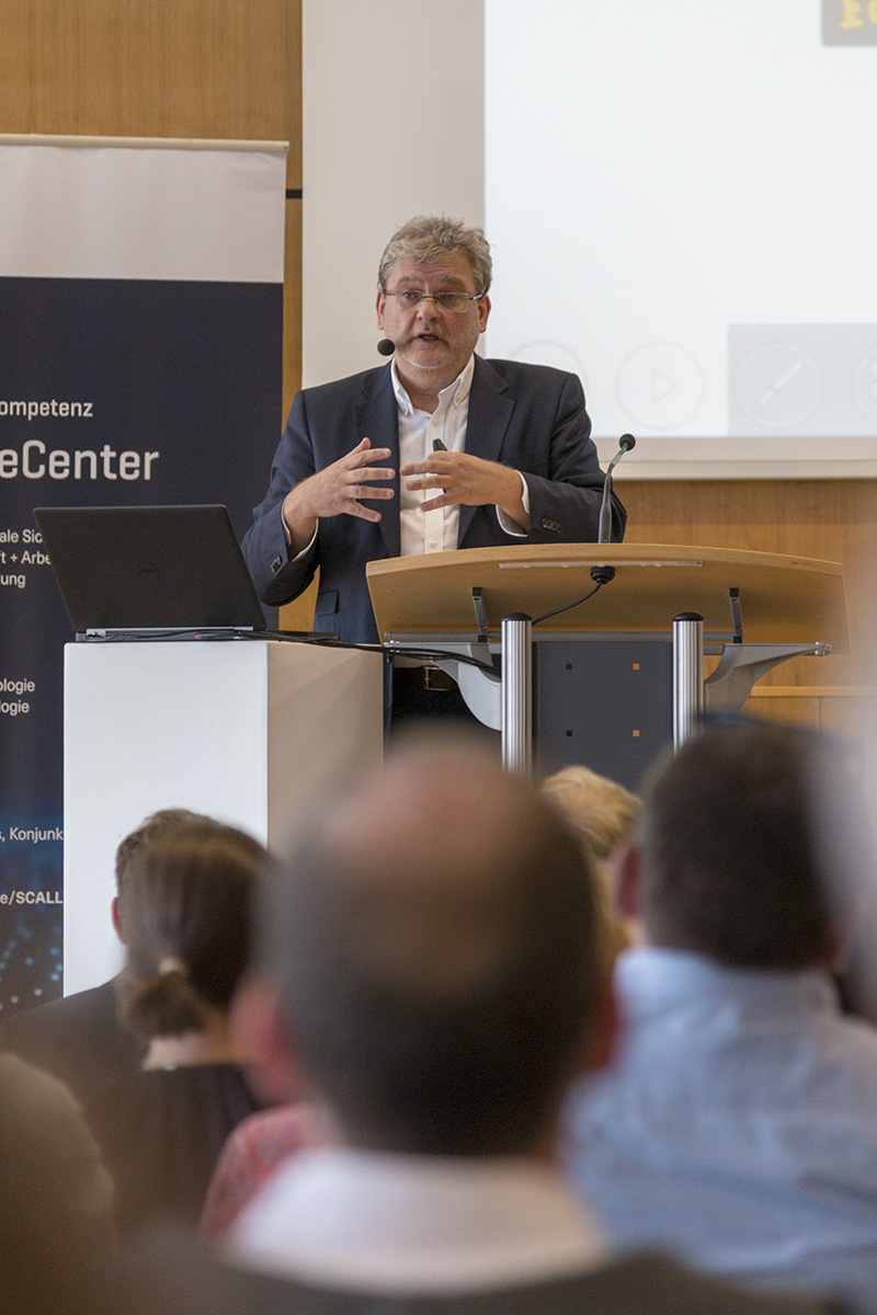 Ein Mann im Anzug steht an einem Podium und hält einen Vortrag. Er vertritt die Hochschule Coburg. Während er spricht, gestikuliert er mit den Händen. Im Hintergrund sind eine Projektionsfläche und ein Banner zu sehen, im Vordergrund sitzt ein aufmerksames Publikum.