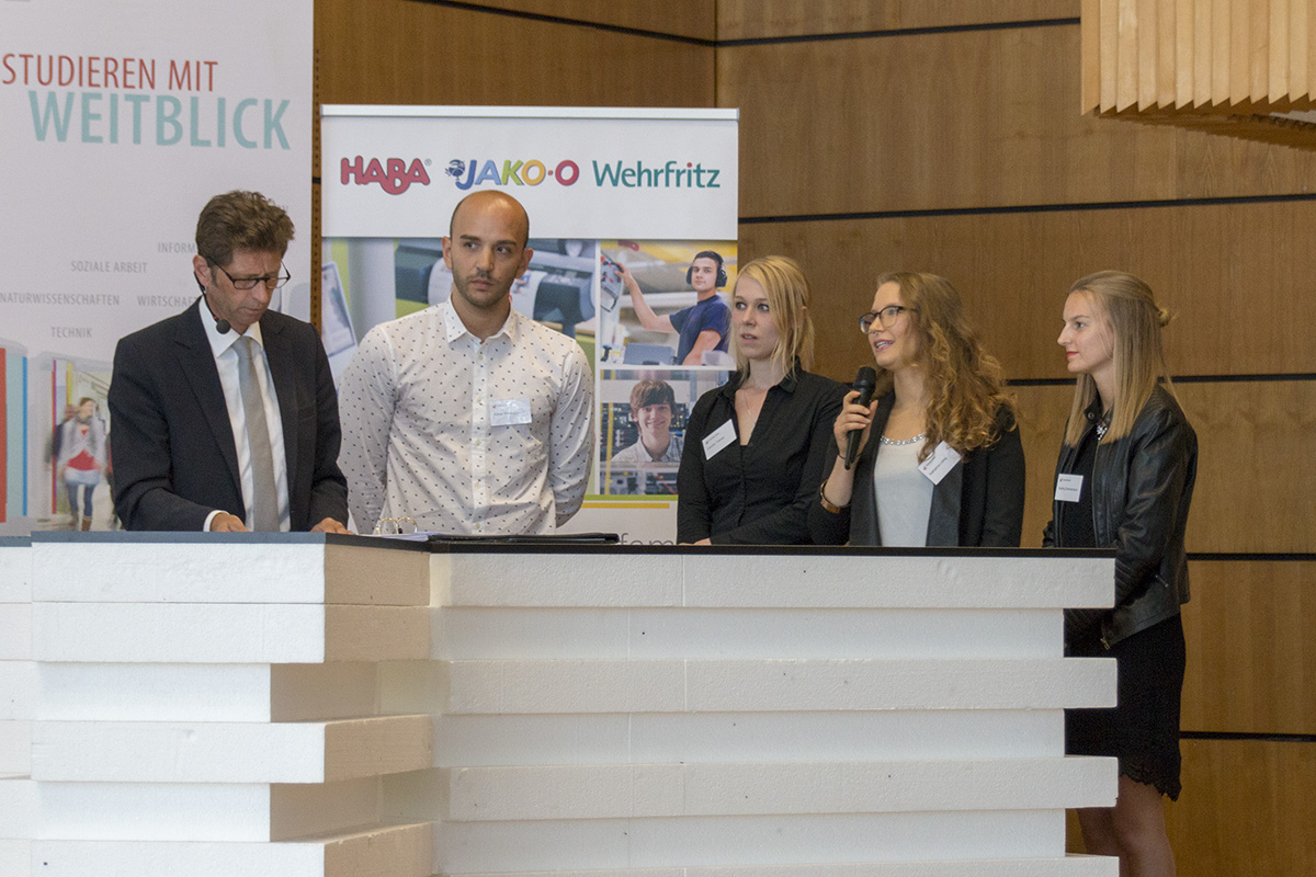 Eine Gruppe von fünf Personen steht hinter einem Podium an der Hochschule Coburg. Ein Mann im Anzug hält Papiere in der Hand, während eine Frau ins Mikrofon spricht. Im Hintergrund ist ein Werbebanner zu sehen, das auf die Bedeutung der Veranstaltung an der renommierten Hochschule hinweist.