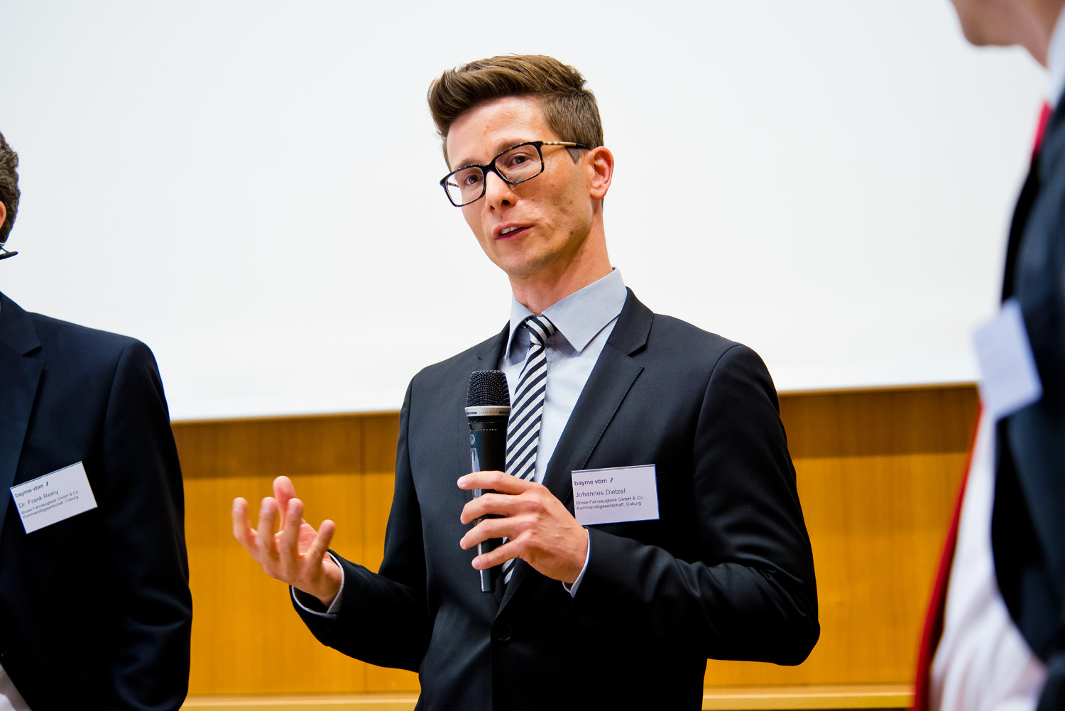 Ein Mann in Anzug und Krawatte spricht in ein Mikrofon und gestikuliert mit einer Hand vor einem hellen Hintergrund. Wahrscheinlich hält er einen Vortrag bei einer Veranstaltung der Hochschule Coburg. Andere Personen in Anzügen sind teilweise sichtbar, was die formelle Atmosphäre des Anlasses verstärkt.