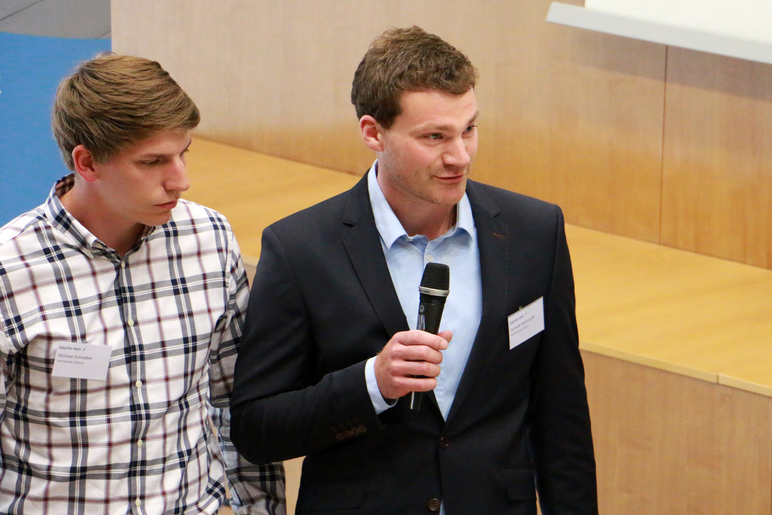 Zwei Männer sprechen auf der Konferenz der Hochschule Coburg. Der Mann links trägt ein kariertes Hemd, der Mann rechts steht im blauen Anzug und mit Mikrofon selbstbewusst vor einem Holzpult.