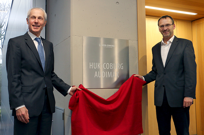 Zwei Männer in Anzügen enthüllen mit einem roten Tuch ein Schild mit der Aufschrift „HUK Coburg Audimax“. Sie stehen zu beiden Seiten des Schildes im Inneren der Hochschule Coburg und lächeln in die Kamera.