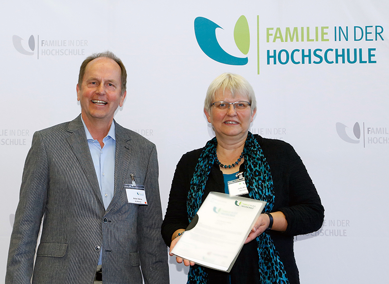 Zwei Personen stehen lächelnd vor einem „Familie in der Hochschule“-Banner. Die Person rechts, Vertreterin der Hochschule Coburg, hält eine Urkunde in der Hand. Beide sind im Business-Outfit gekleidet.