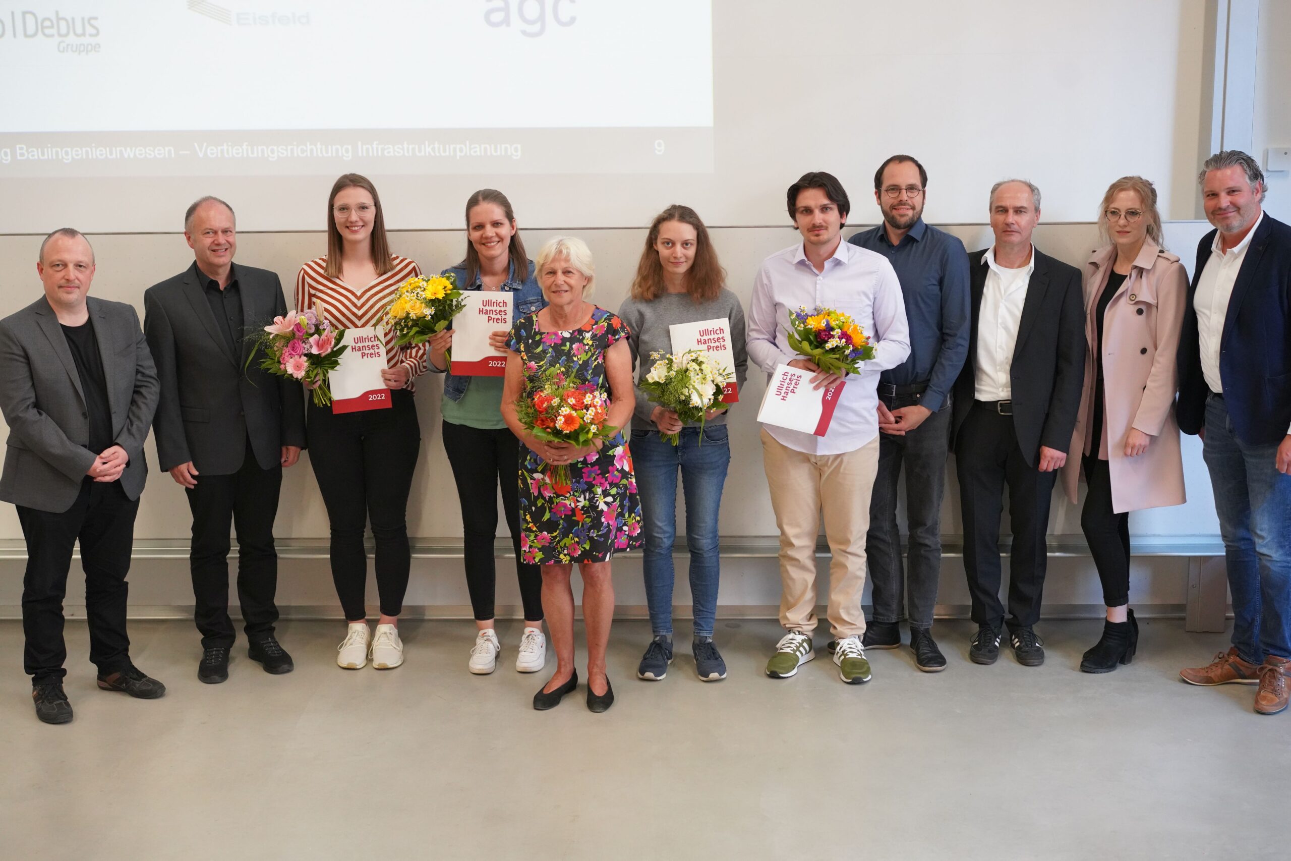 Eine Gruppe von Menschen, einige mit Zertifikaten und Blumensträußen in den Händen, steht in einer Reihe vor einer weißen Tafel an der Hochschule Coburg. Sie sind leger und formell gekleidet und lächeln in die Kamera.