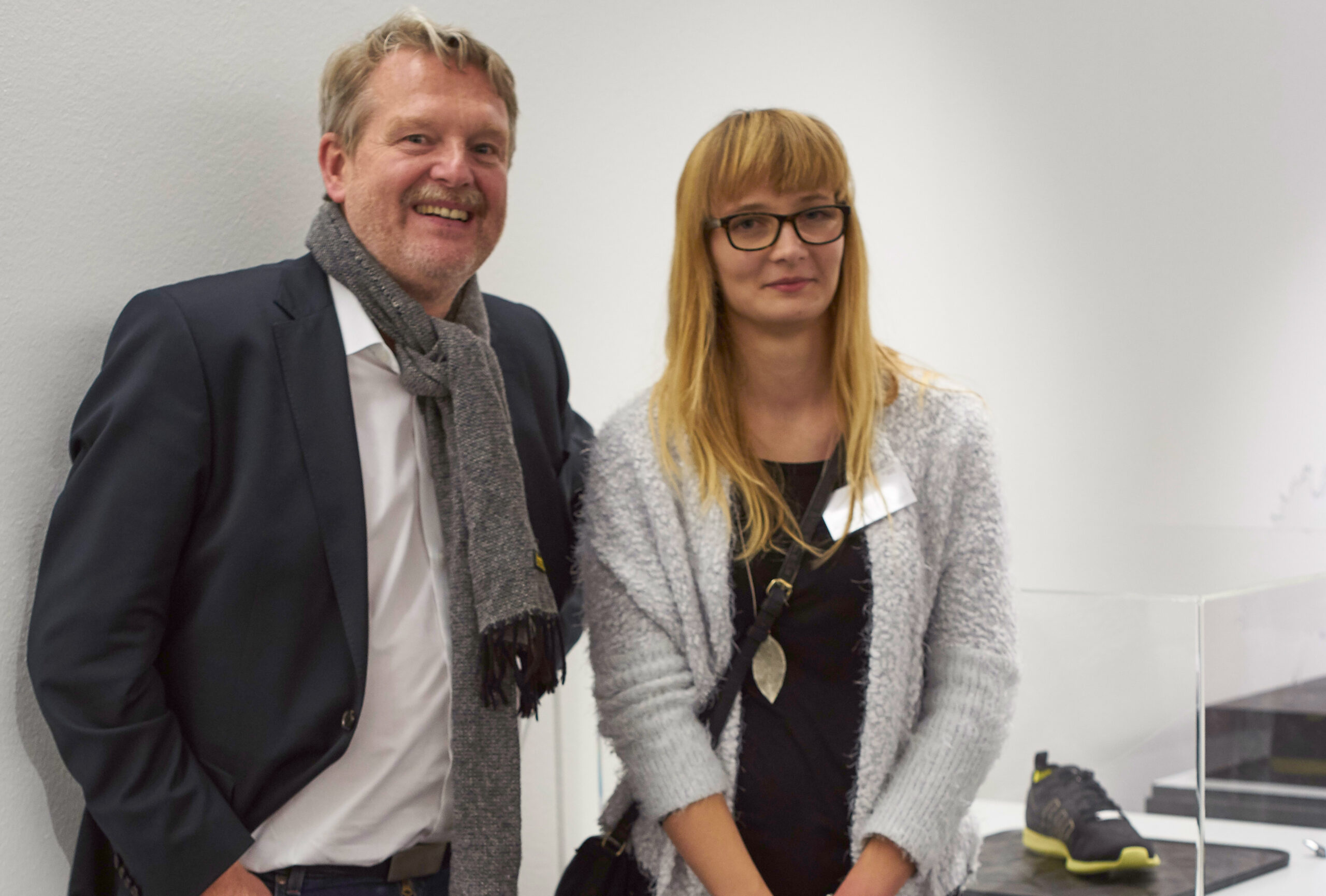 Ein Mann und eine Frau stehen zusammen in einem Raum der Hochschule Coburg. Der Mann, Anzug und Schal tragend, lächelt herzlich, während die Frau mit Brille und heller Strickjacke neutral wirkt. Im Hintergrund ist ein Turnschuh in einer Vitrine ausgestellt.