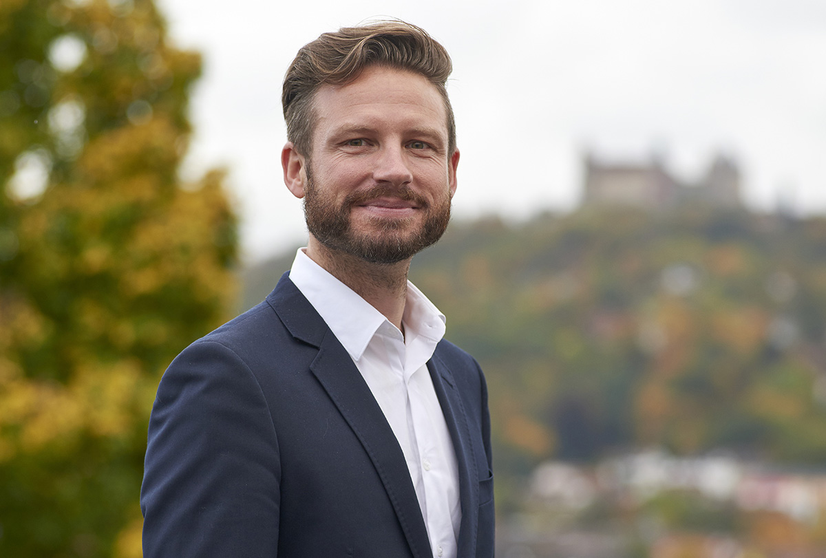 Ein Mann mit Bart und gestyltem Haar trägt einen marineblauen Blazer und ein weißes Hemd und steht im Freien. Hinter ihm sind im verschwommenen Hintergrund Bäume und ein entfernter Hügel zu sehen, auf dem sich das repräsentative Gebäude der Hochschule Coburg erhebt.