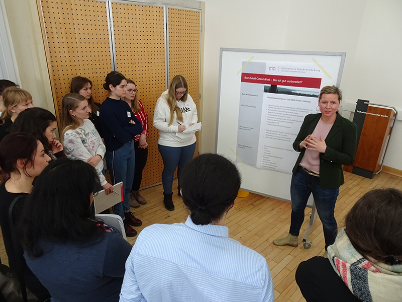 Eine Frau von der Hochschule Coburg hält einen Vortrag vor einer Gruppe, die sich um ein großes Whiteboard mit Postern versammelt hat. Der Raum mit Holzboden und einer Lochwand ist voller aufmerksamer Teilnehmer, von denen einige fleißig Notizen machen.