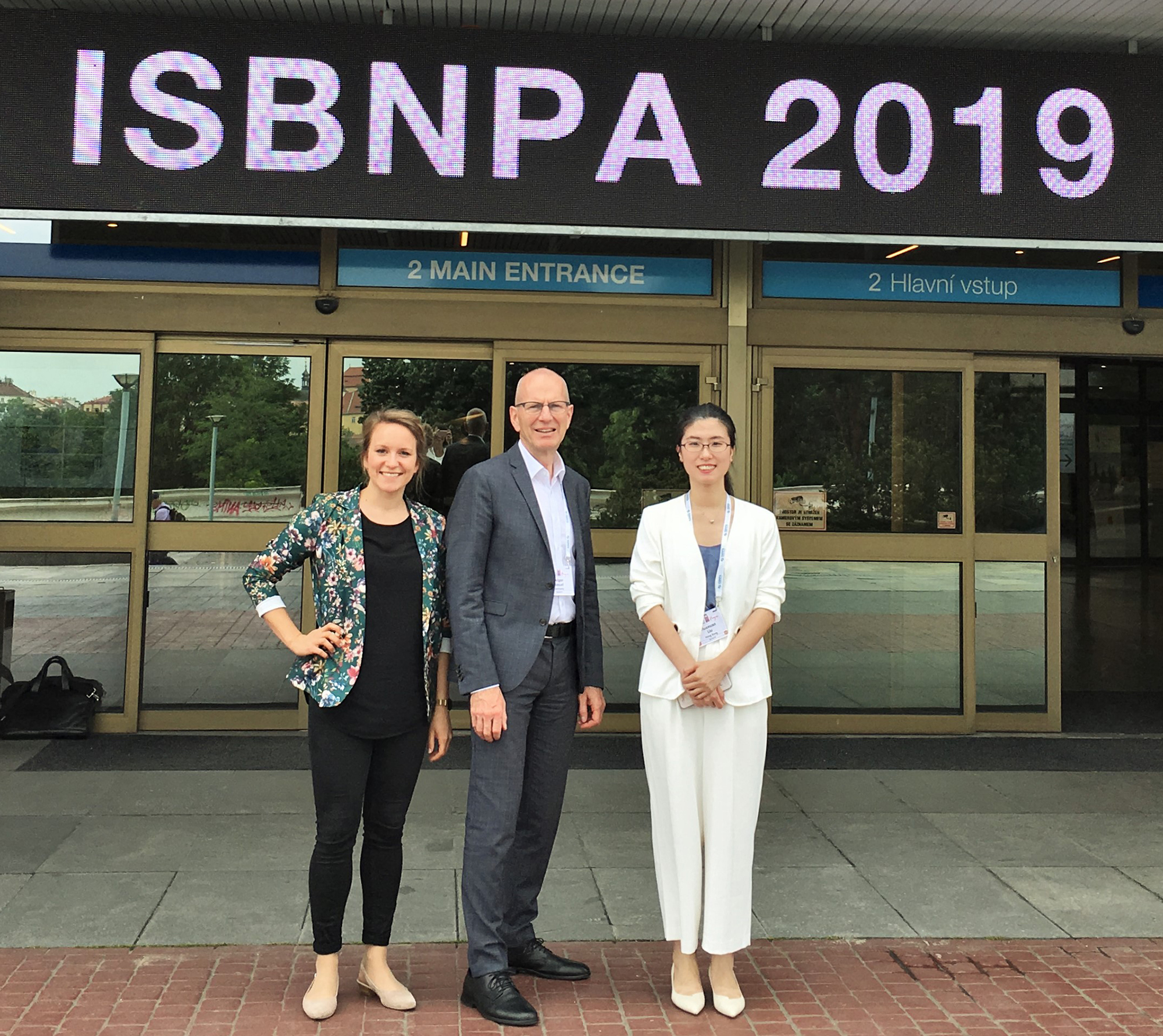 Drei Menschen stehen lächelnd vor einem Gebäudeeingang, über dem ein Schild mit der Aufschrift „ISBNPA 2019“ hängt. Flankiert von den beiden Frauen steht der Mann stolz im Business-Outfit und macht ein unvergessliches Gruppenfoto an der Hochschule Coburg.