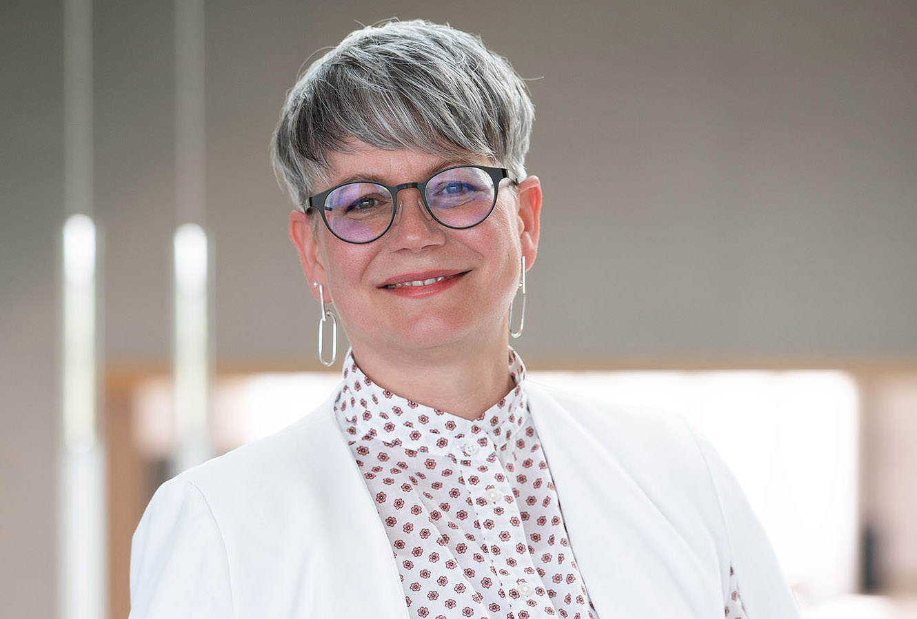 Eine Person mit kurzen grauen Haaren, Brille, weißer Jacke und gemustertem Hemd mit dem Wappen der Hochschule Coburg lächelt sanft. Der Hintergrund ist leicht verschwommen, wodurch eine neutrale Atmosphäre entsteht.