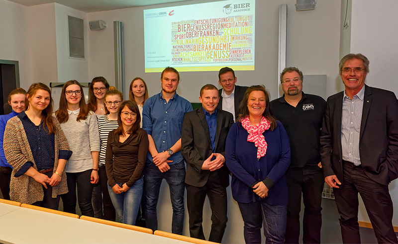 Eine Gruppe von Menschen steht in einem Hörsaal der Hochschule Coburg vor einer Projektionsfläche, auf der ein bunter Text angezeigt wird. Sie lächeln und sind in Freizeit- oder Businesskleidung gekleidet, als würden sie an einem spannenden Workshop oder einer Präsentation teilnehmen.