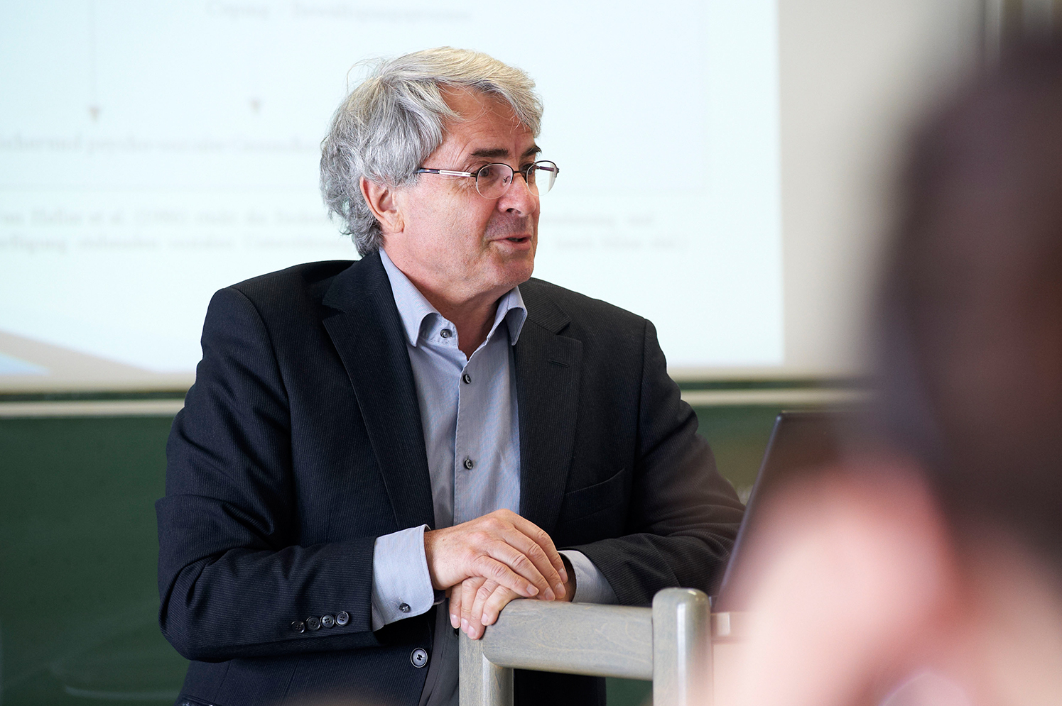 Ein älterer Mann mit grauen Haaren und Brille trägt einen schwarzen Anzug und ein hellblaues Hemd und steht an einem Podium vor einer Präsentationsleinwand der Hochschule Coburg.