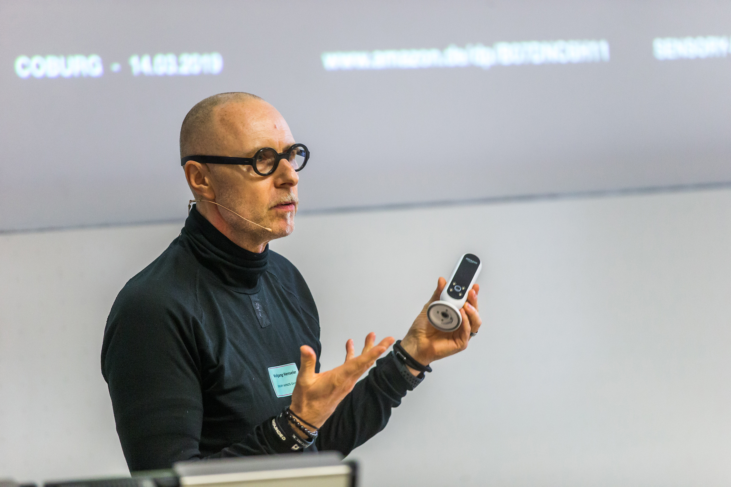 Eine Person mit Brille und Headset-Mikrofon hält einen Vortrag an der Hochschule Coburg. Sie hält eine Fernbedienung in der Hand und gestikuliert lebhaft, während im Hintergrund auf einem unscharfen Bildschirm Text angezeigt wird.