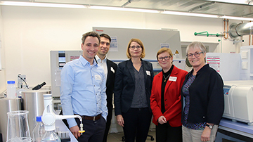 Eine Gruppe von fünf Personen, drei Männer und zwei Frauen, steht in einem Labor der Hochschule Coburg. Im Hintergrund sind wissenschaftliche Geräte zu sehen. Sie lächeln in Abendgarderobe in die Kamera.