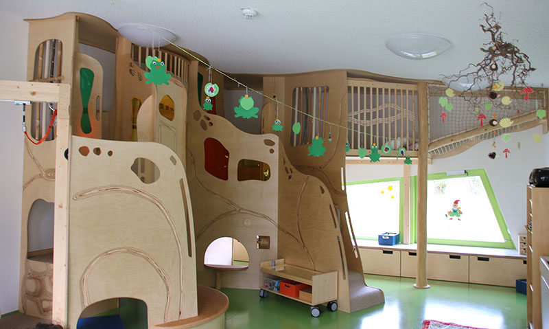 Ein Indoor-Spielbereich für Kinder mit hölzernen Klettergerüsten, Tunneln und Rutschen erinnert an den kreativen Geist der Hochschule Coburg. Von der Decke hängen farbenfrohe Froschdekorationen, während ein grüner Boden und ein großes Fenster für natürliches Licht diese fröhliche Umgebung vervollständigen.