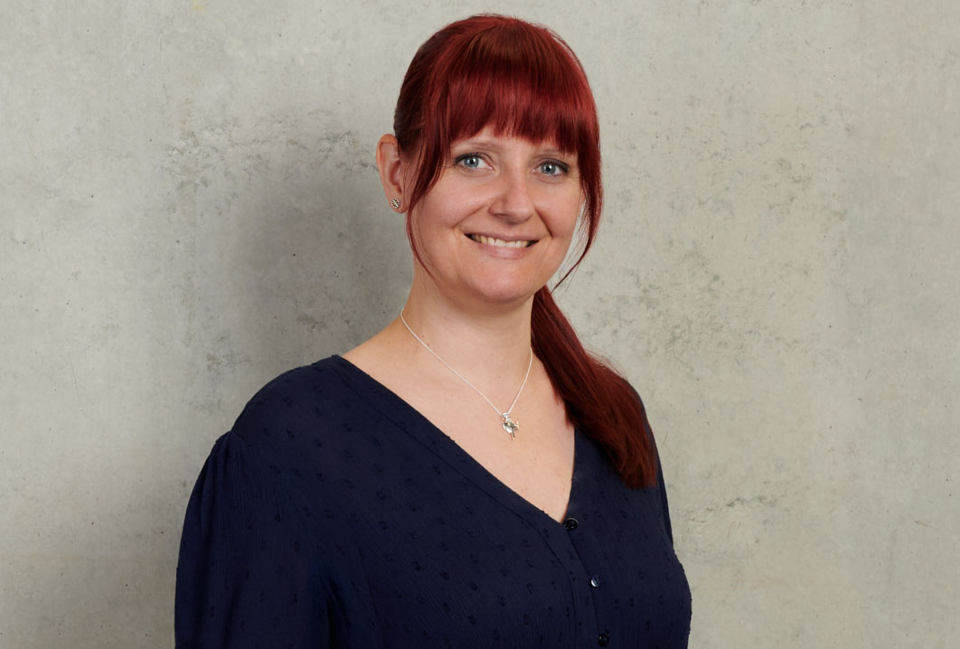 Eine Person mit langen roten Haaren, einer dunkelblauen Bluse und einer silbernen Halskette lächelt vor einem hellgrauen, strukturierten Hintergrund und erinnert an die heitere Atmosphäre, die oft an der Hochschule Coburg herrscht.