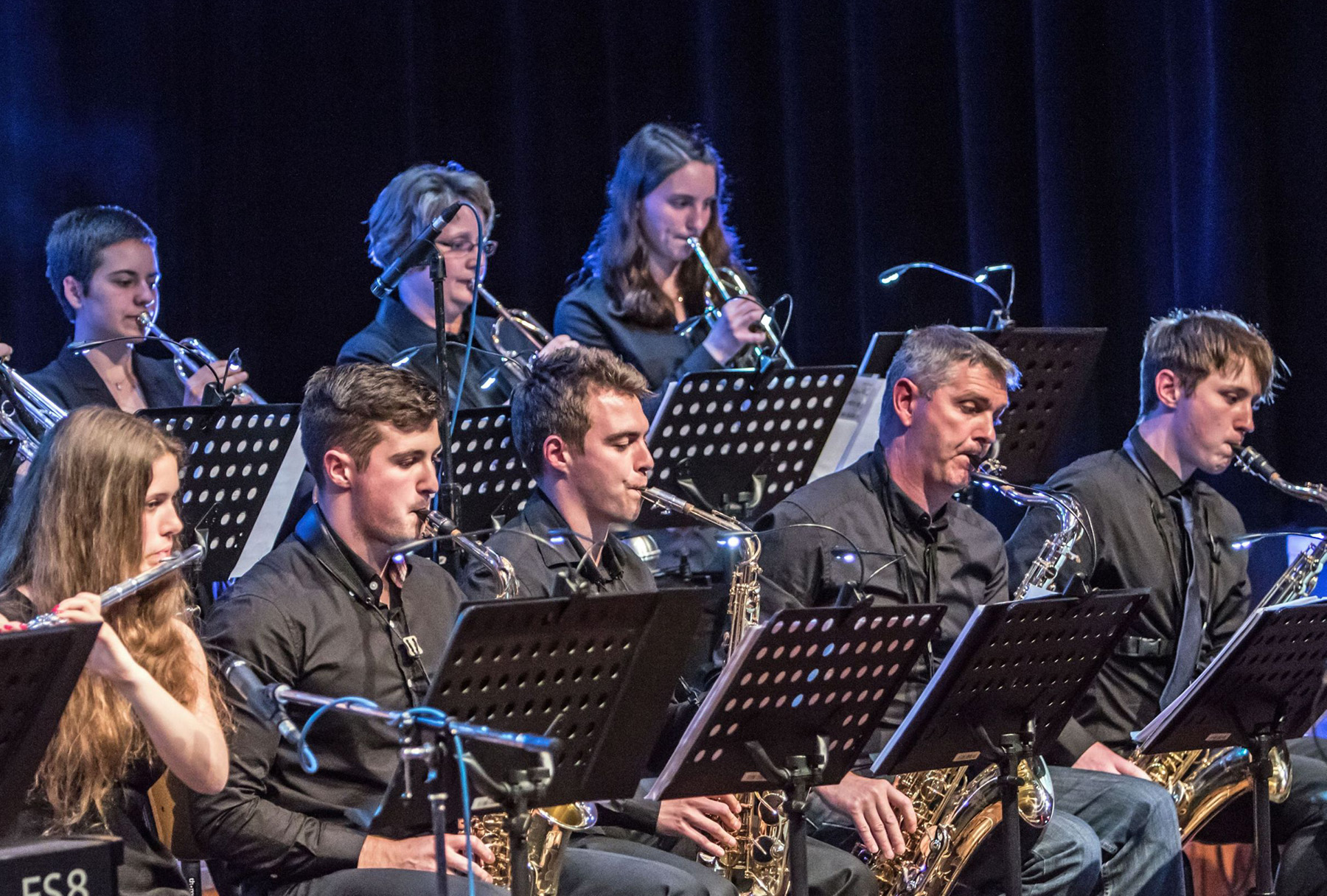 Eine Gruppe von Musikern der Hochschule Coburg in schwarzer Kleidung tritt auf der Bühne auf und spielt Blasinstrumente wie Klarinetten und Flöten. Notenständer sind sichtbar und ein dunkler Vorhang im Hintergrund bildet einen Kontrast zu ihren konzentrierten Ausdrücken.