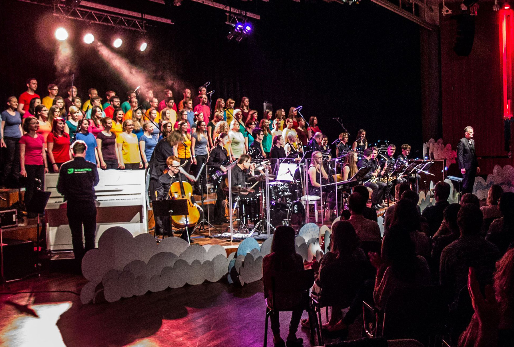 Ein Chor und ein Orchester der Hochschule Coburg treten unter buntem Licht auf der Bühne auf. Der Chor, in leuchtende Hemden gekleidet, steht hinter Musikern, die verschiedene Instrumente spielen. Das Publikum verfolgt die lebhafte Aufführung in einem schwach beleuchteten Auditorium.