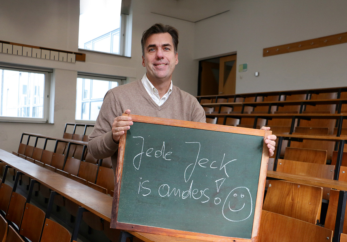 Eine Person steht in einem Hörsaal der Hochschule Coburg und hält eine Tafel mit dem Spruch „Jede Jeck ist anders!“ hoch. In dem leeren Saal stehen Reihen von Holztischen und Sitzgelegenheiten.