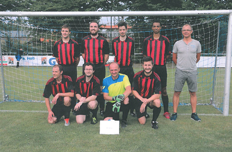 Eine Gruppe von acht Männern in rot-schwarzen Fußballtrikots der Hochschule Coburg posiert vor einem Tor auf einem Rasenplatz. Sieben Spieler tragen die Teamfarben, einer trägt Freizeitkleidung. Vor ihnen steht ein kleines Schild, im Hintergrund sind Bäume und ein Gebäude zu sehen.