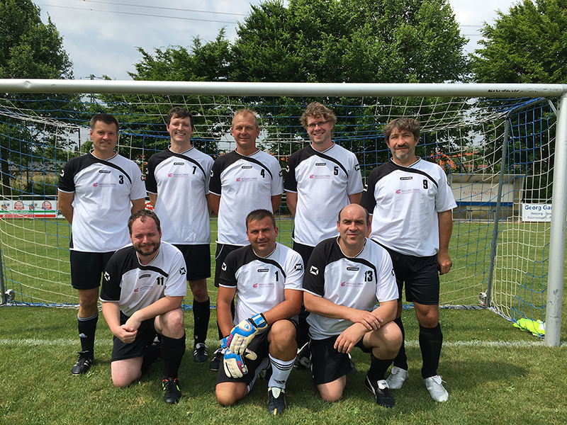 Eine Gruppe von acht Männern in weiß-schwarzen Fußballtrikots der Hochschule Coburg posiert für ein Mannschaftsfoto vor einem Tor auf einer Wiese. Sie lächeln, einige stehen, andere knien. Im Hintergrund sind Bäume und ein Teilblick auf ein Stadion zu sehen.