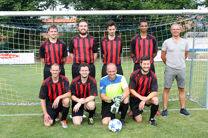 Eine Fußballmannschaft der Hochschule Coburg posiert auf dem Feld. Acht Männer stehen oder knien vor einem Tor. Sie tragen rot-schwarz gestreifte Trikots und schwarze Hosen. Der Torwart, in Uniform und Handschuhen, hält einen Fußball. Bäume und ein Zaun bilden eine malerische Kulisse.