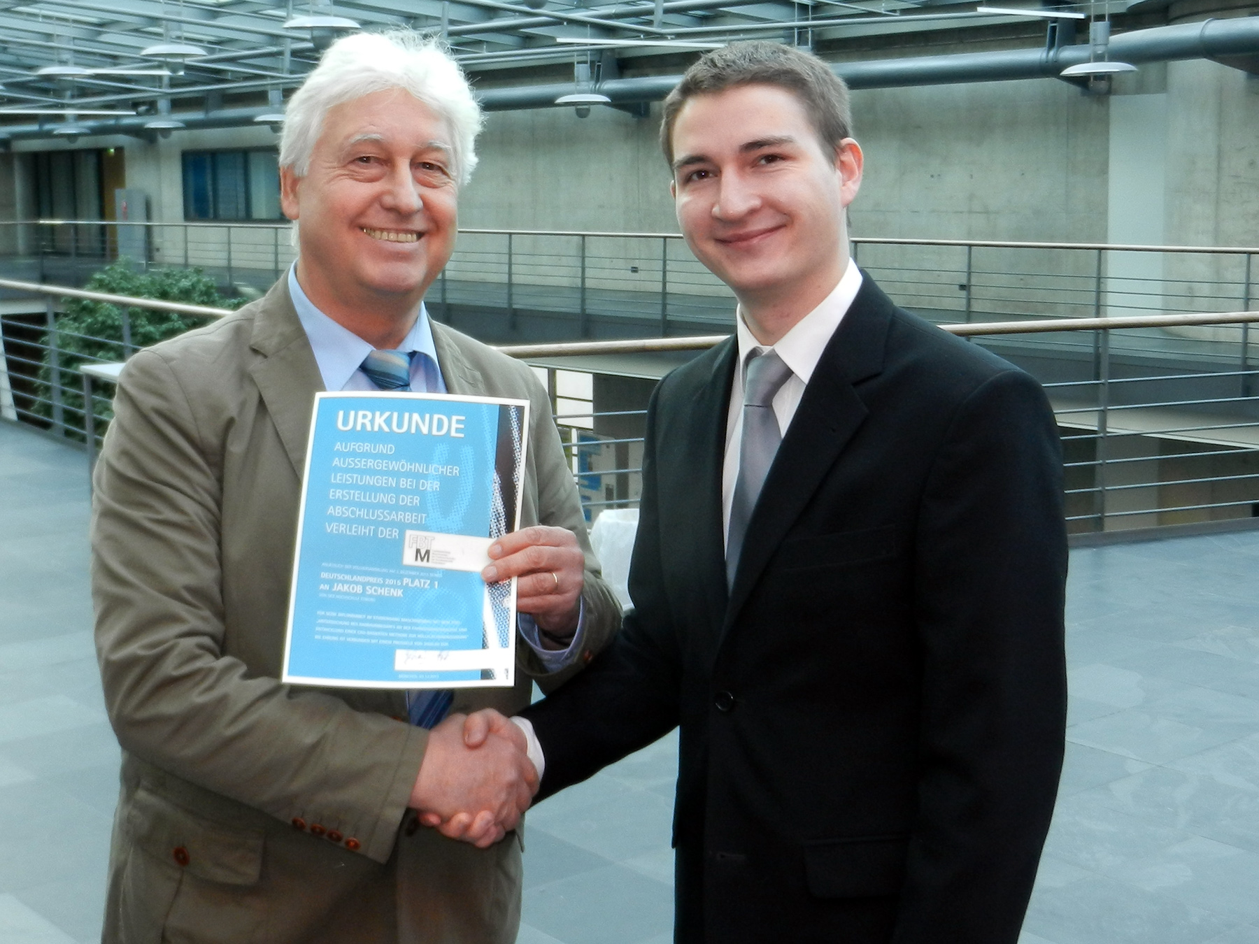 Zwei Männer in Anzügen schütteln sich die Hände, während einer von ihnen ein Zertifikat mit der Aufschrift „Urkunde“ in der Hand hält. Sie stehen in einem modernen Innenraum, der repräsentativ für die Hochschule Coburg ist, mit einem Metallgeländer und Pflanzen im Hintergrund.