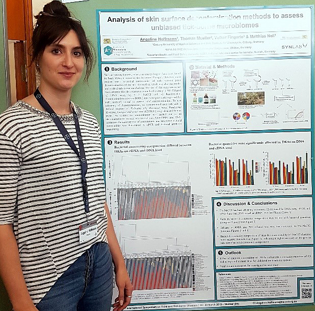 Eine Person steht neben einem wissenschaftlichen Poster der Hochschule Coburg über die Analyse von Methoden zur Dekontamination von Hautoberflächen auf durch Zecken übertragene Mikrobiome. Das Poster enthält Grafiken, Text und Bilder. Die Person trägt ein Abzeichen und ein gestreiftes Hemd.