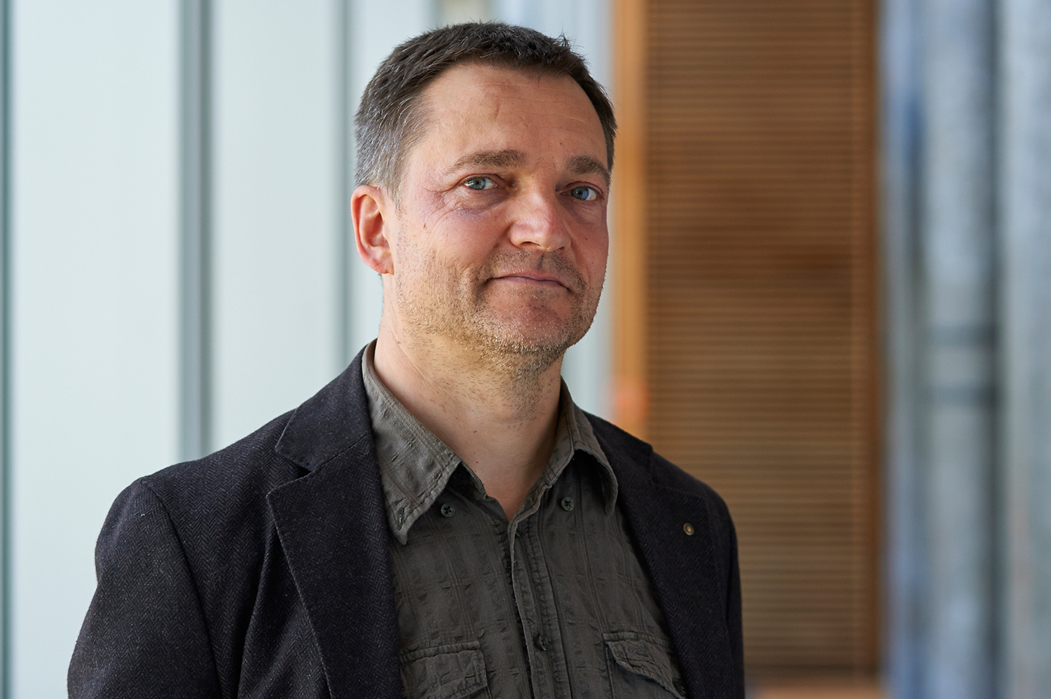 Ein Mann in einem dunklen Blazer und einem grauen Hemd steht im Innenbereich der Hochschule Coburg. Der Hintergrund besteht aus vertikalen Paneelen und einem Holzabschnitt, was ihm ein modernes und minimalistisches Aussehen verleiht.