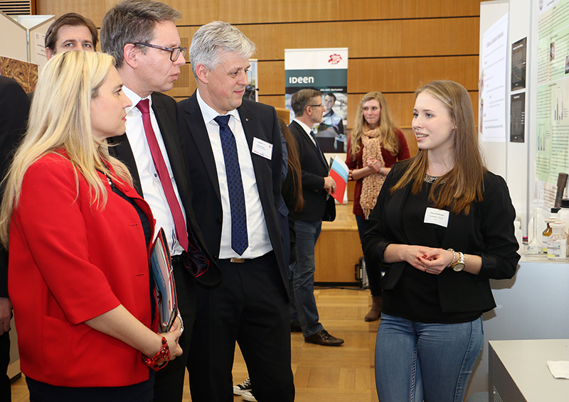 Bei einer öffentlichen Veranstaltung der Hochschule Coburg präsentiert eine junge Frau ihre Arbeit vor drei Erwachsenen, zwei Männern in Anzügen und einer Frau in einer roten Jacke. Im Hintergrund sind Poster und Banner zu sehen, die den lebendigen Geist der Wissenschaft widerspiegeln. Überall im Raum sind weitere Teilnehmer zu sehen.