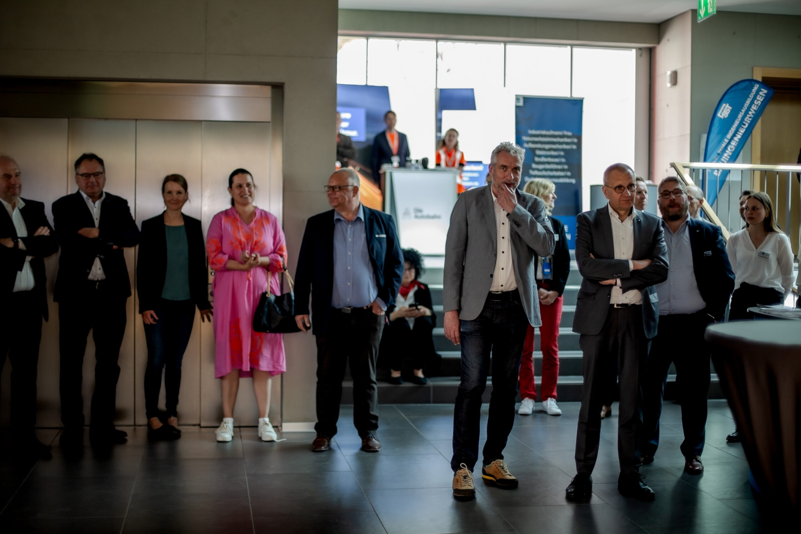 Eine Gruppe von Menschen in Business-Kleidung steht in einem modernen Foyer der Hochschule Coburg. Sie beobachten aufmerksam etwas, das außerhalb ihres Blickfelds liegt. Einige stehen mit verschränkten Armen da, andere haben die Hände in den Taschen. Im Hintergrund sind ein Empfangstresen und Aufzugstüren zu sehen.