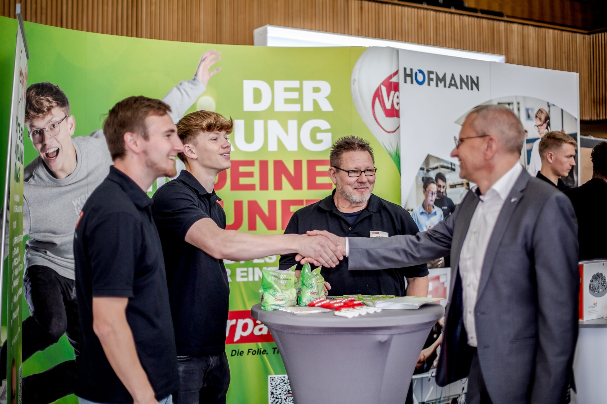 Vier Männer stehen an einem Messestand um einen mit Snacks und Werbematerial beladenen Tisch. Zwei Männer links schütteln einem Mann im Anzug rechts die Hand, während ein Mann mit Brille aufmerksam zusieht. Bunte Banner, die für die Hochschule Coburg werben, dienen als lebendiger Hintergrund.