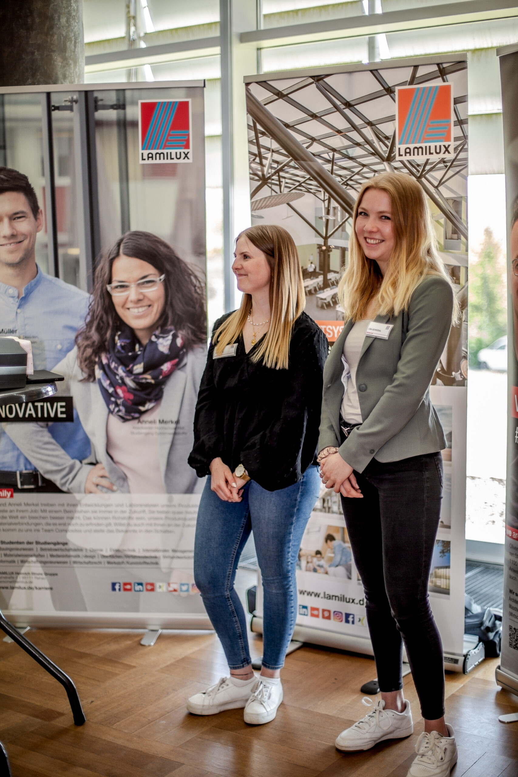 Zwei Frauen stehen lächelnd vor LamLUX-Plakaten an der Hochschule Coburg. Die Plakate zeigen Bilder von Menschen und architektonischen Strukturen. Lässig gekleidet und mit Namensschildern versehen stehen sie auf einem Holzboden in einem gut beleuchteten Raum.