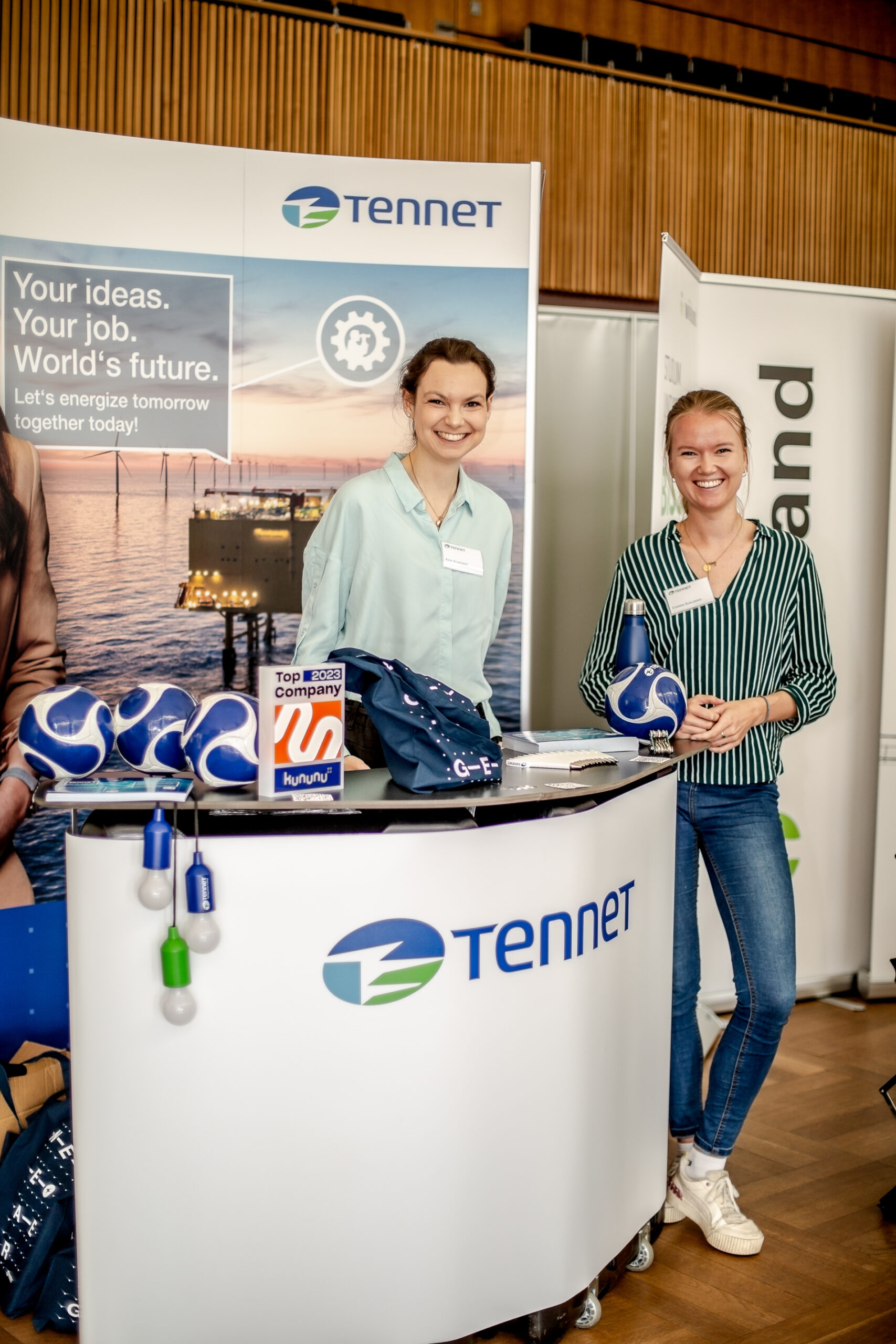 Zwei Menschen lächeln an einem Stand von TenneT, an dem ein Banner mit dem Slogan „Deine Ideen. Dein Job. Die Zukunft der Welt“ hängt. Der Stand, der in der Hochschule Coburg präsentiert wird, bietet Fußbälle mit Team-Branding und Werbeartikel in einer modernen Inneneinrichtung.