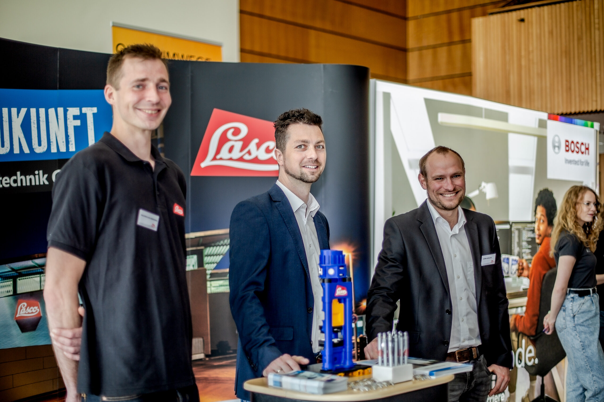 Drei Männer in Businesskleidung stehen an einem Messestand mit Lasco-Logo und einem Banner der Hochschule Coburg. Lächelnd stehen sie hinter einem Tisch, auf dem Industriegeräte und Broschüren ausgestellt sind. Im Hintergrund ist ein Bosch-Logo zu sehen.