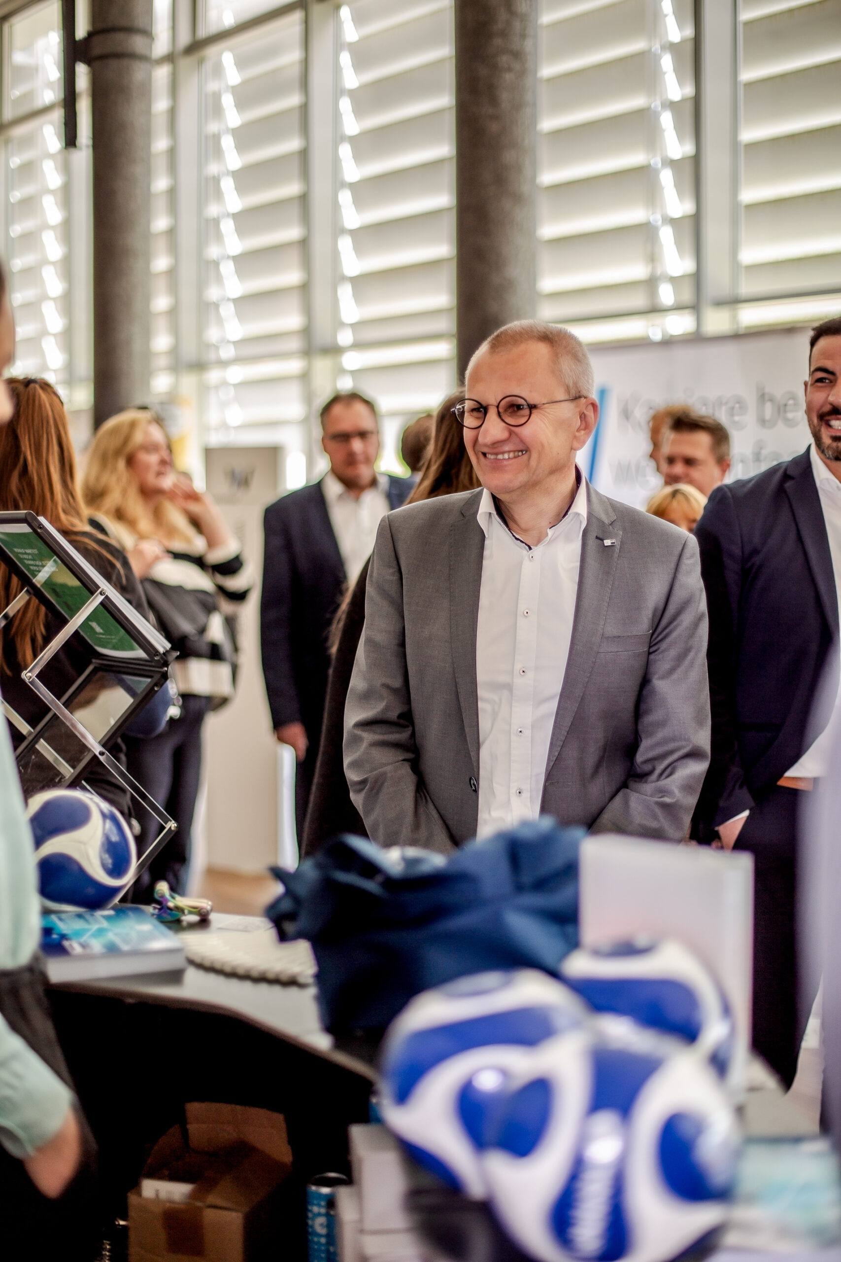 Ein Mann im grauen Anzug, der die Hochschule Coburg vertritt, steht lächelnd inmitten einer Gruppe von Menschen im Innenbereich. Im Vordergrund stehen Tische mit blau-weißen Fußbällen und Werbeartikeln. Der Raum hat große Fenster mit teilweise geschlossenen Jalousien.