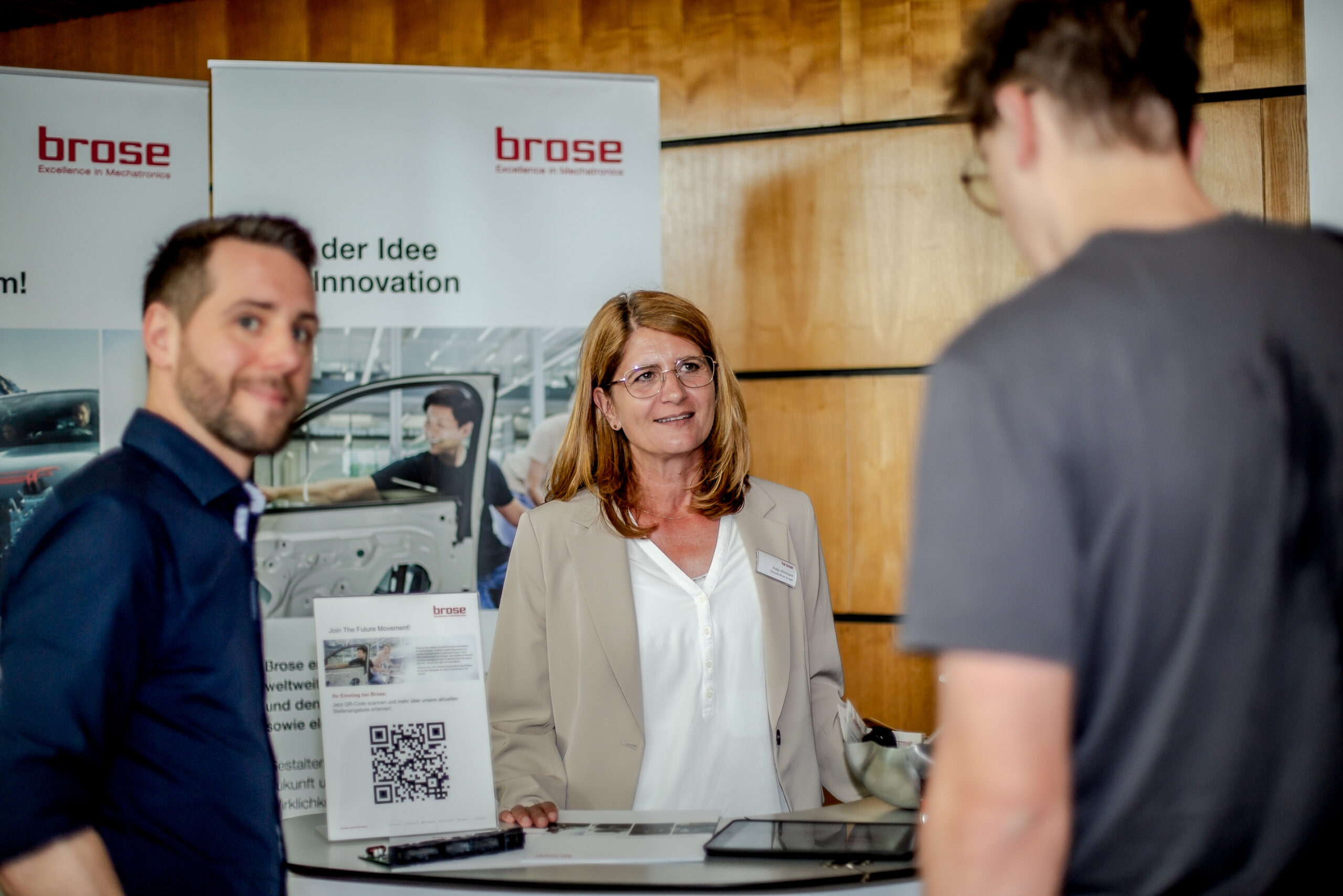 Eine Frau und zwei Männer unterhalten sich an einem Stand mit dem „brose“-Branding der Hochschule Coburg im Hintergrund. Das Ambiente ist mit Holzvertäfelung, einer Broschüre und einem QR-Code ausgestattet.